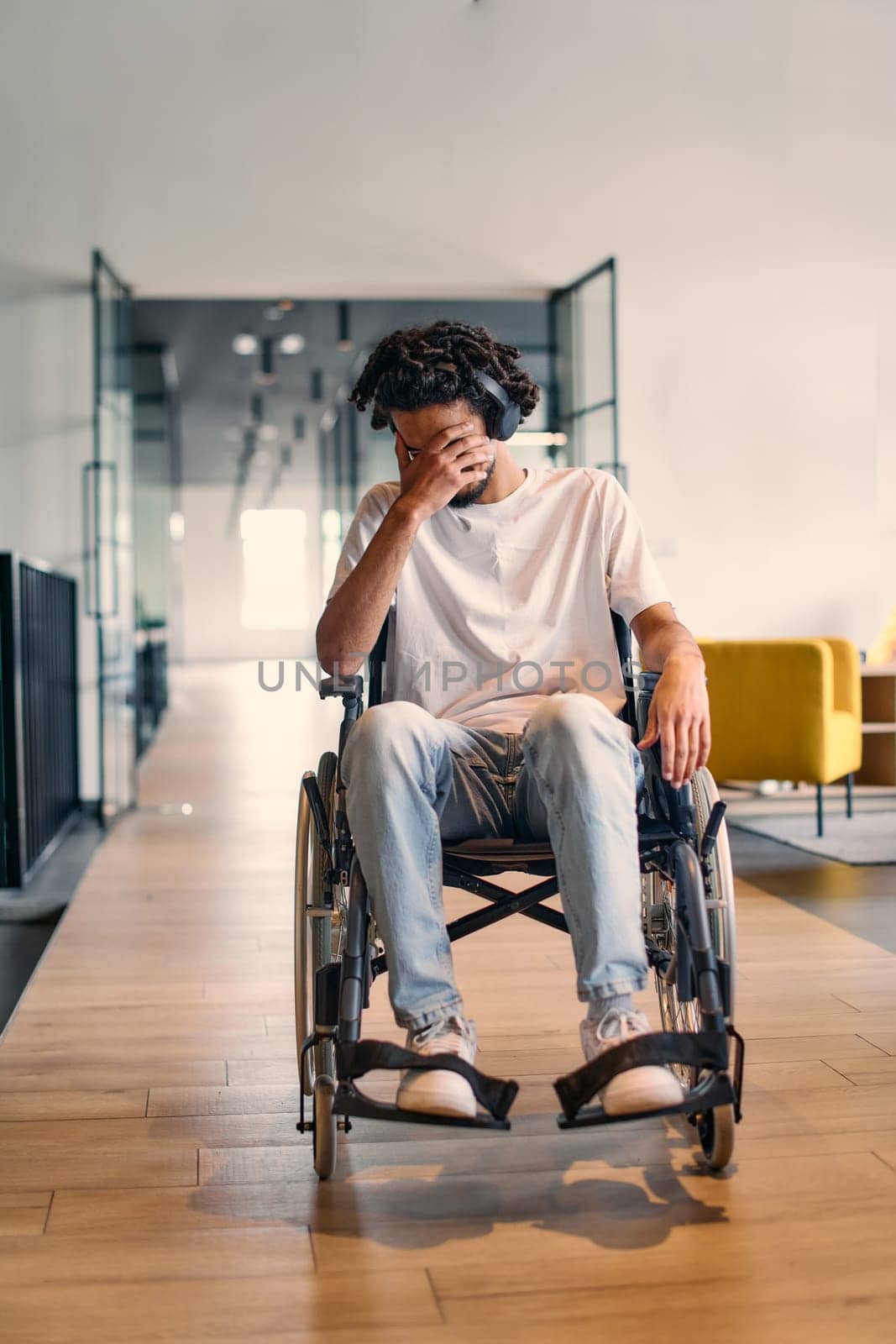 A African-American teenager in a wheelchair sits sadly amidst the bustling backdrop of a modern startup office, surrounded by his business colleagues