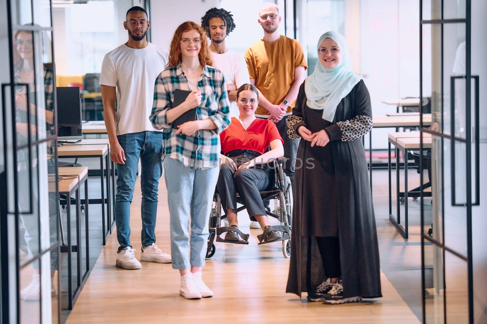 A diverse group of young business people walking a corridor in the glass-enclosed office of a modern startup, including a person in a wheelchair and a woman wearing a hijab by dotshock