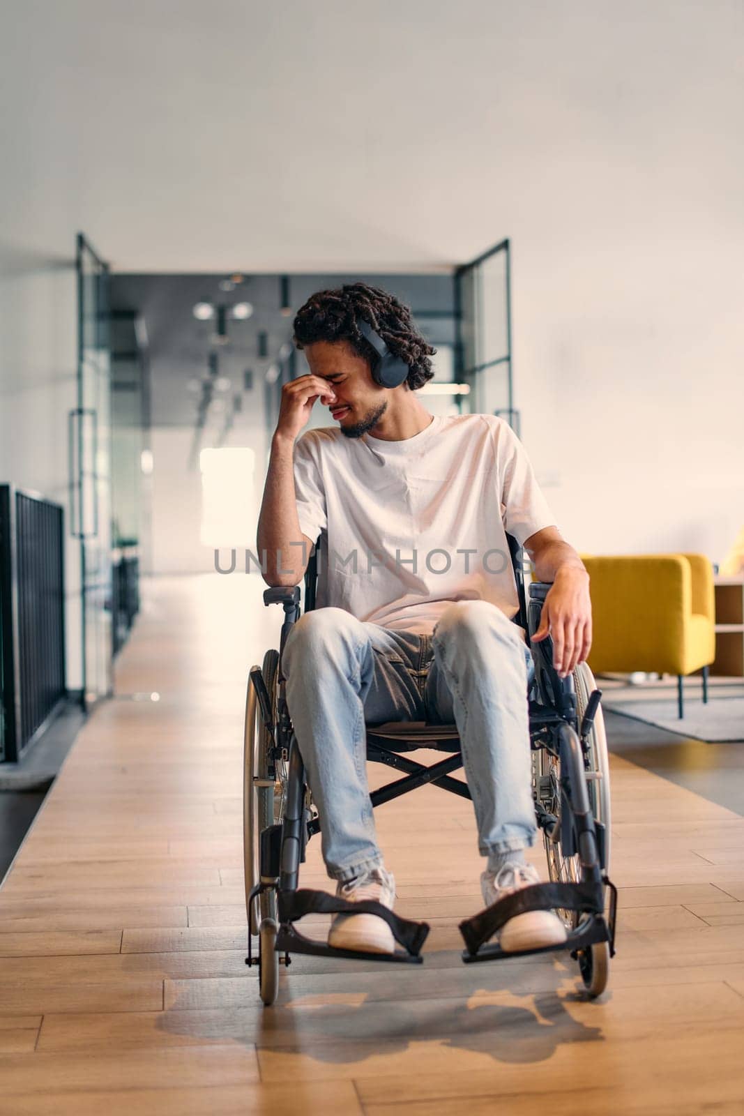 A African-American teenager in a wheelchair sits sadly amidst the bustling backdrop of a modern startup office, surrounded by his business colleagues. by dotshock