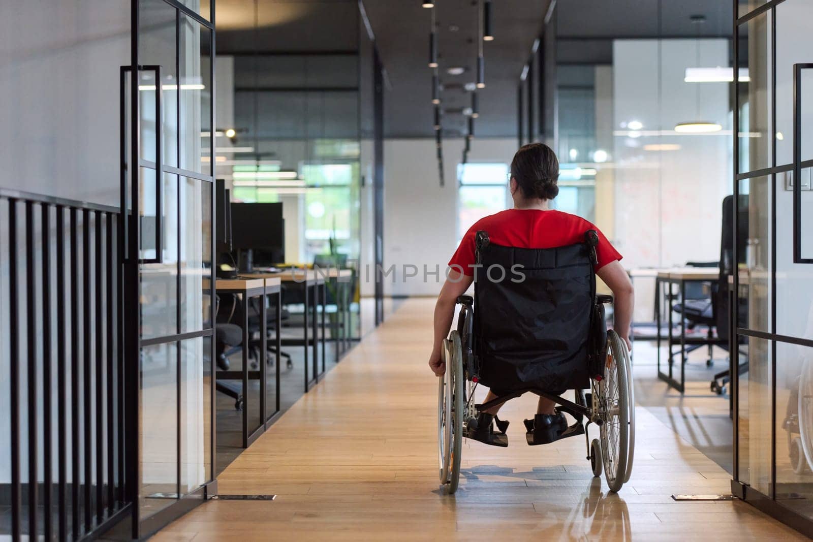 A modern young businesswoman in a wheelchair is surrounded by an inclusive workspace with glass-walled offices, embodying determination and innovation in the business world.