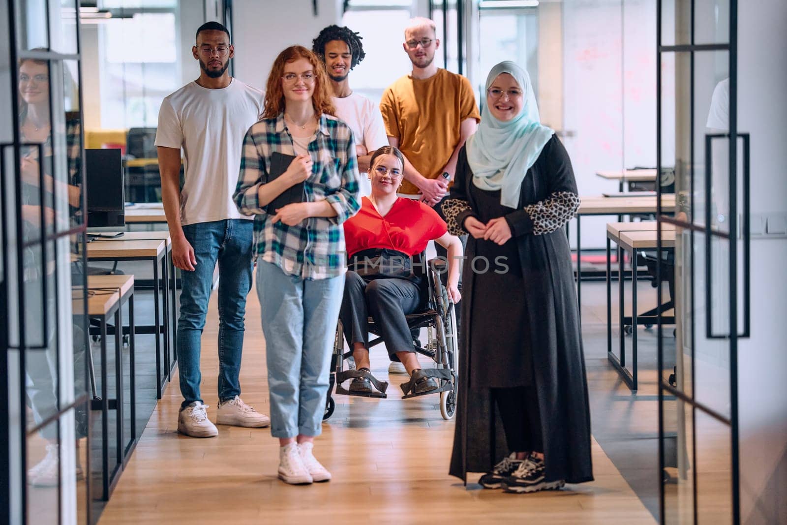 A diverse group of young business people walking a corridor in the glass-enclosed office of a modern startup, including a person in a wheelchair and a woman wearing a hijab by dotshock