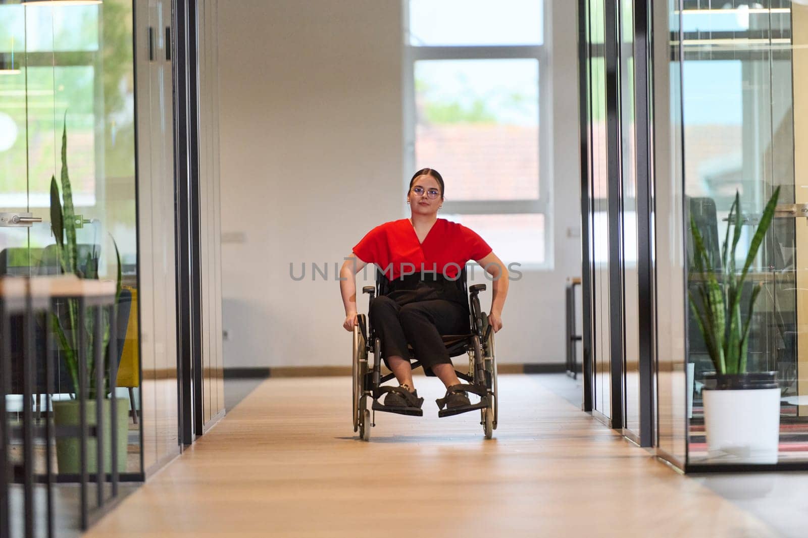 A modern young businesswoman in a wheelchair is surrounded by an inclusive workspace with glass-walled offices, embodying determination and innovation in the business world.