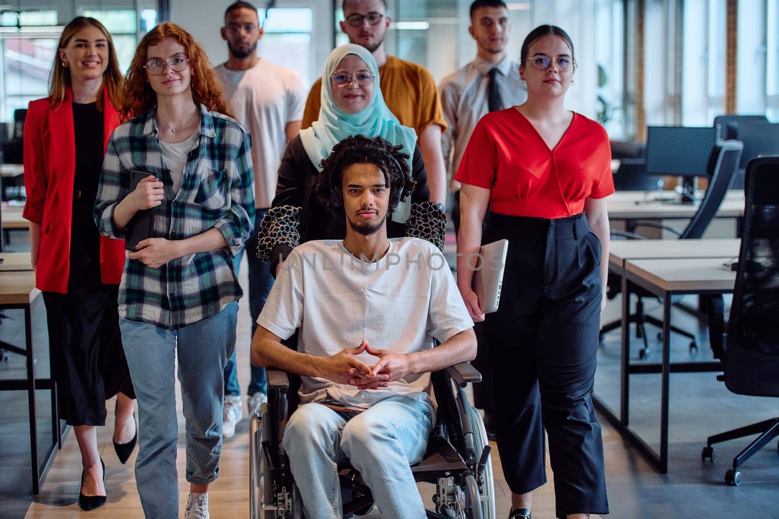 A diverse group of young business people congregates within a modern startup's glass-enclosed office, featuring inclusivity with a person in a wheelchair, an African American young man, and a hijab muslim woman