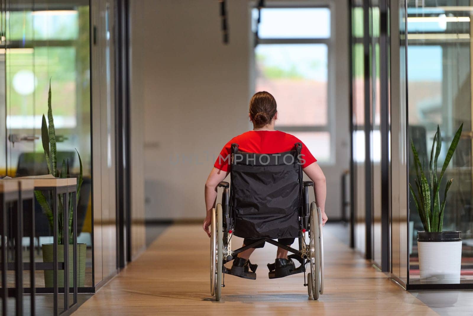 A modern young businesswoman in a wheelchair is surrounded by an inclusive workspace with glass-walled offices, embodying determination and innovation in the business world.