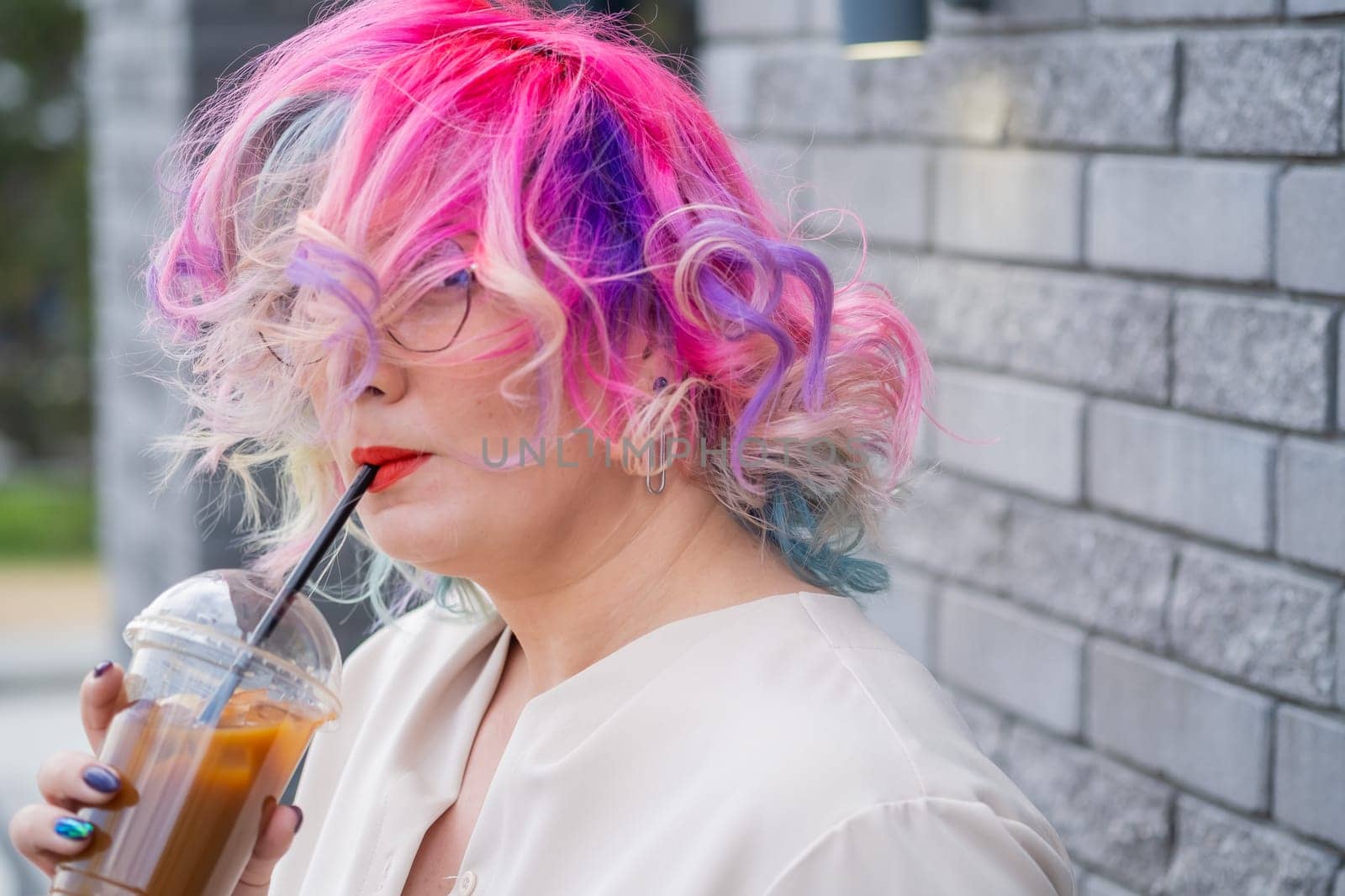 Close-up portrait of curly Caucasian woman with multi-colored hair wearing glasses. The hairstyle model is drinking a cold drink.