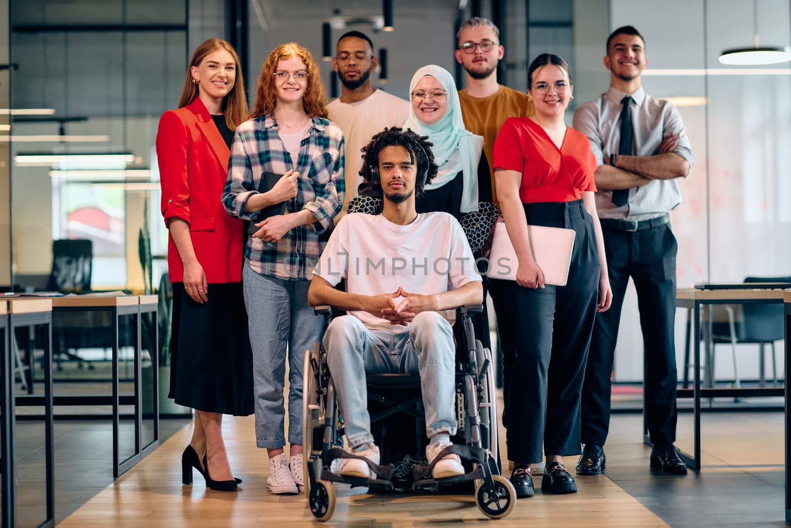 A diverse group of young business people walking a corridor in the glass-enclosed office of a modern startup, including a person in a wheelchair and a woman wearing a hijab.