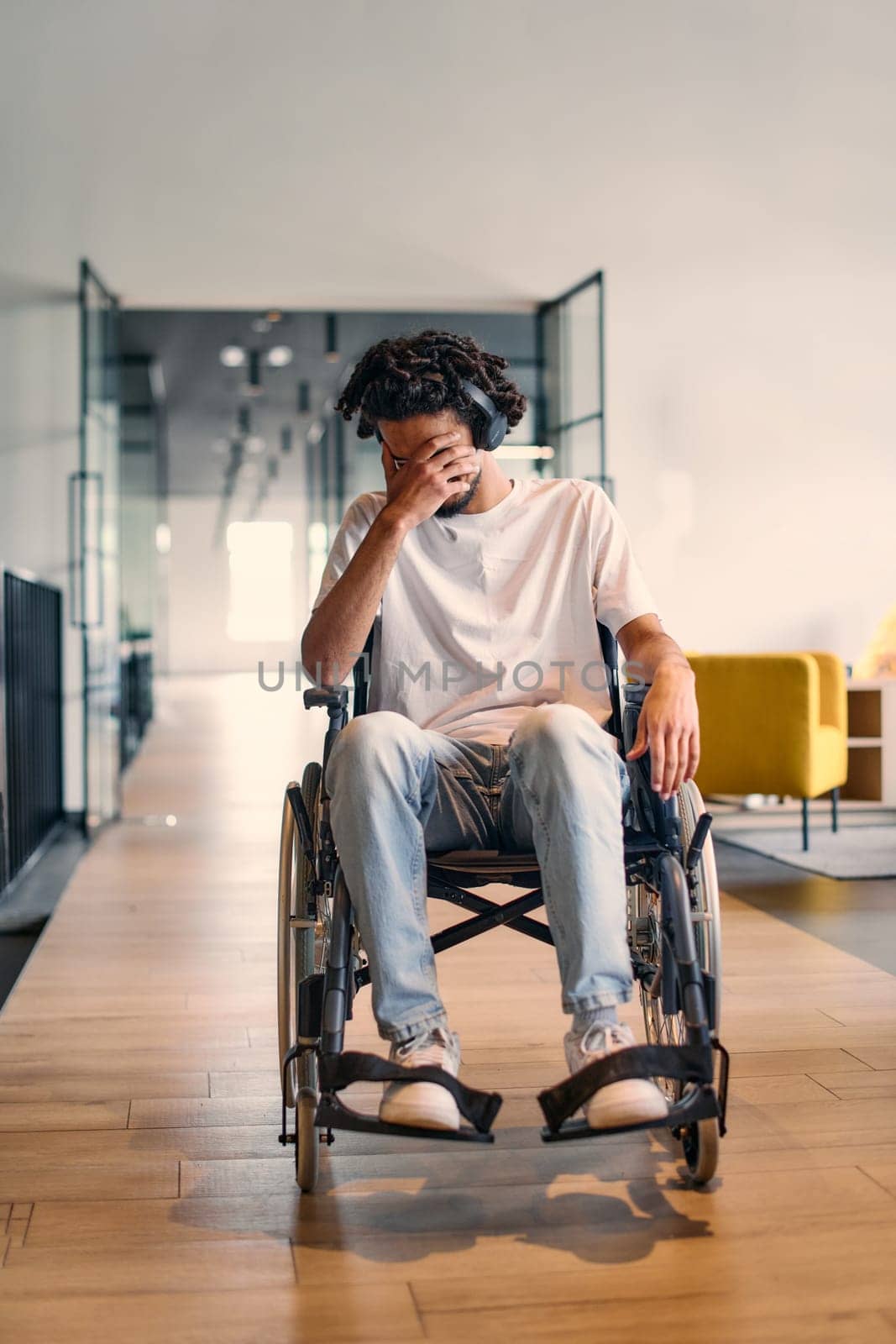 A African-American teenager in a wheelchair sits sadly amidst the bustling backdrop of a modern startup office, surrounded by his business colleagues