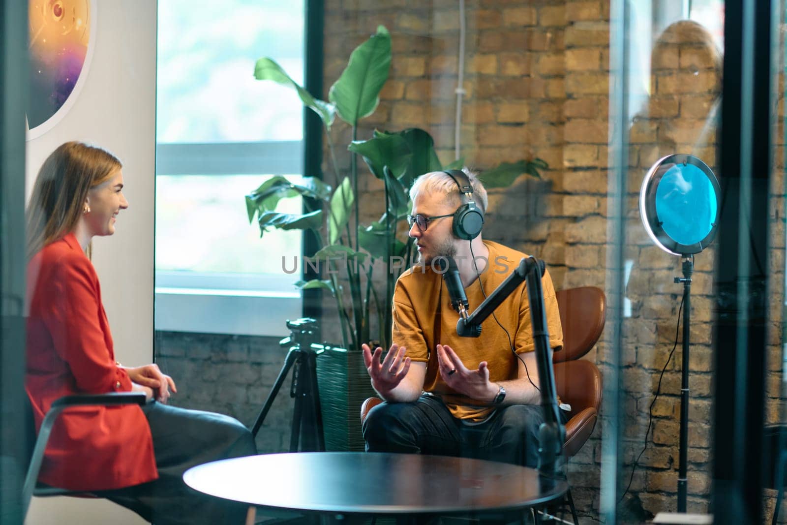 A gathering of young business professionals, some seated in a glass-walled office, engage in a lively conversation and record an online podcast, embodying modern collaboration and dynamic interaction by dotshock