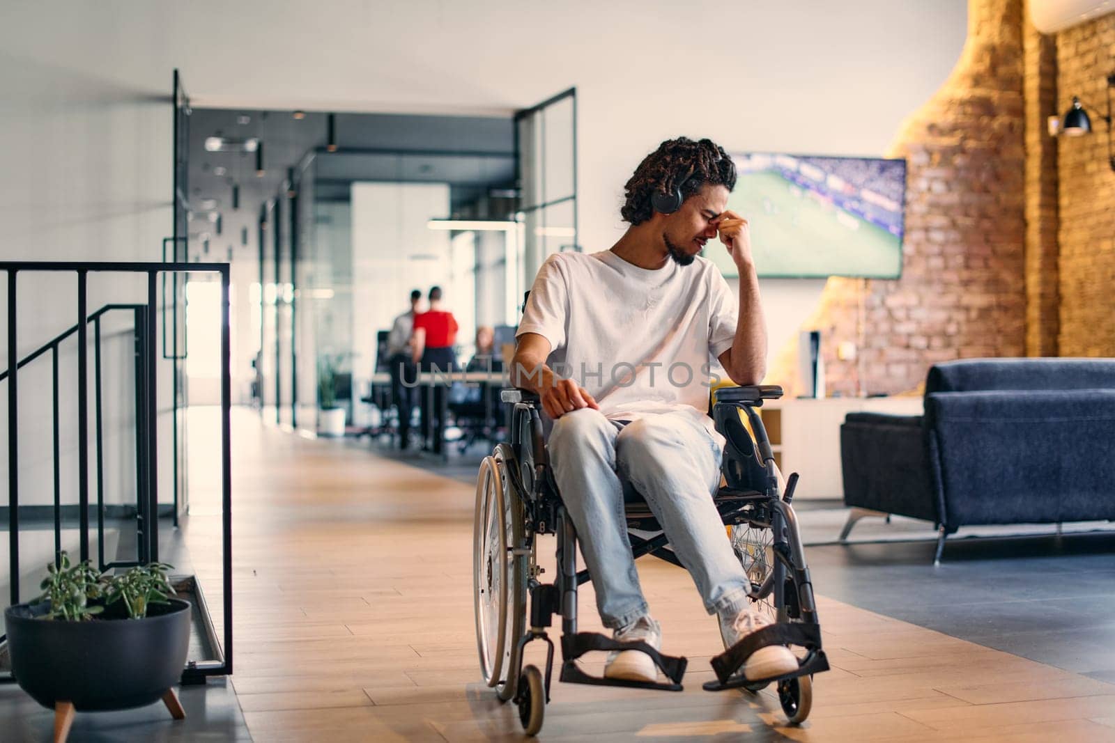 A African-American teenager in a wheelchair sits sadly amidst the bustling backdrop of a modern startup office, surrounded by his business colleagues. by dotshock
