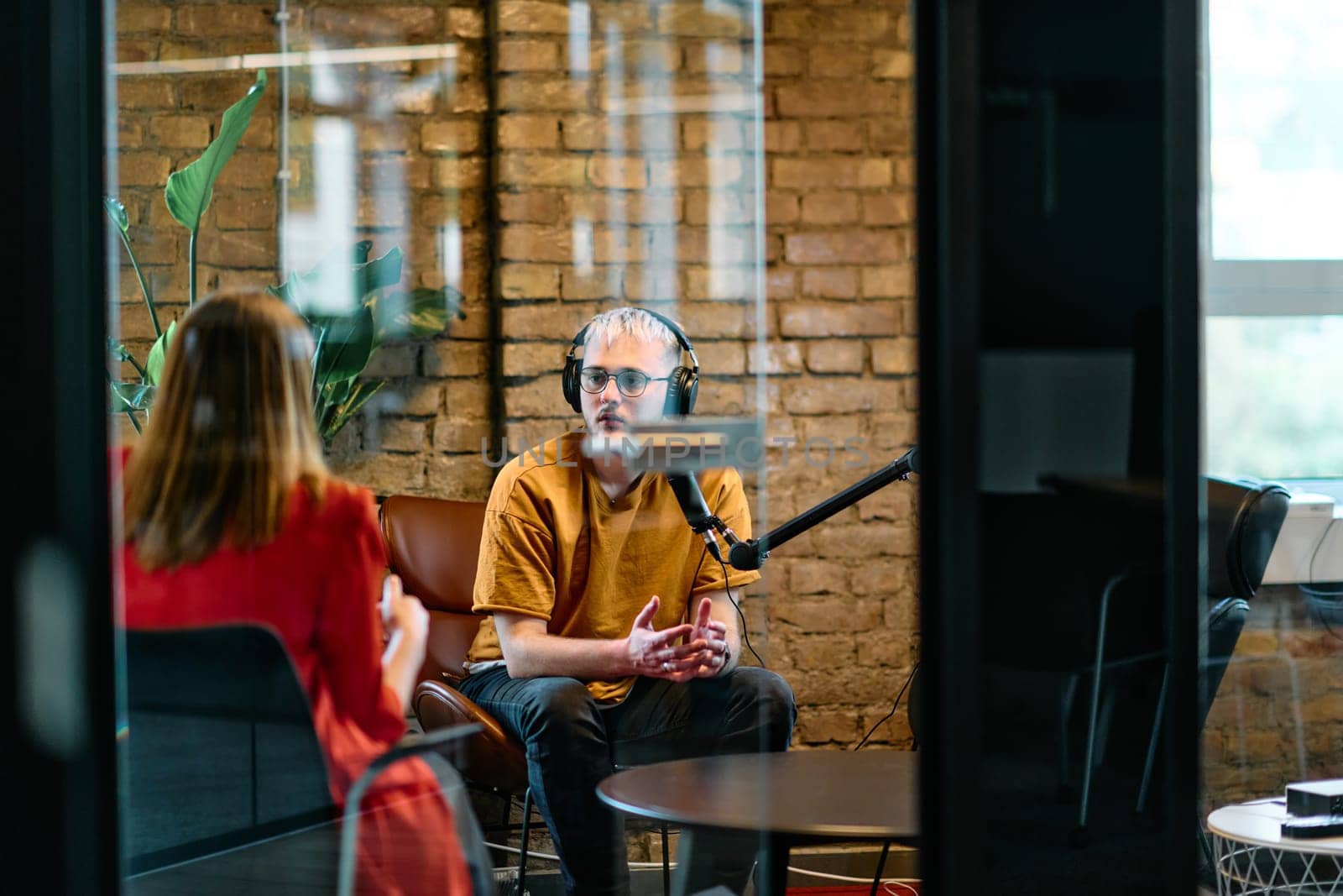 A gathering of young business professionals, some seated in a glass-walled office, engage in a lively conversation and record an online podcast, embodying modern collaboration and dynamic interaction.