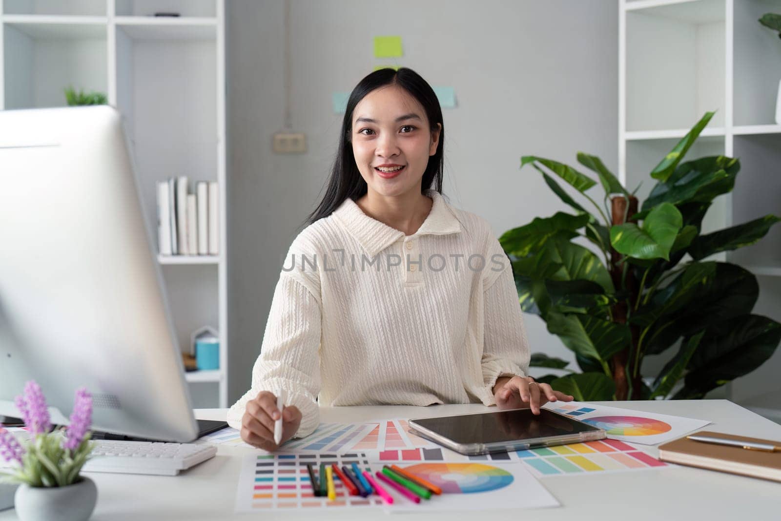 Asian woman freelance graphic designer working with color swatch samples and computer at desk in home office, young lady choosing color gamma for new design project.