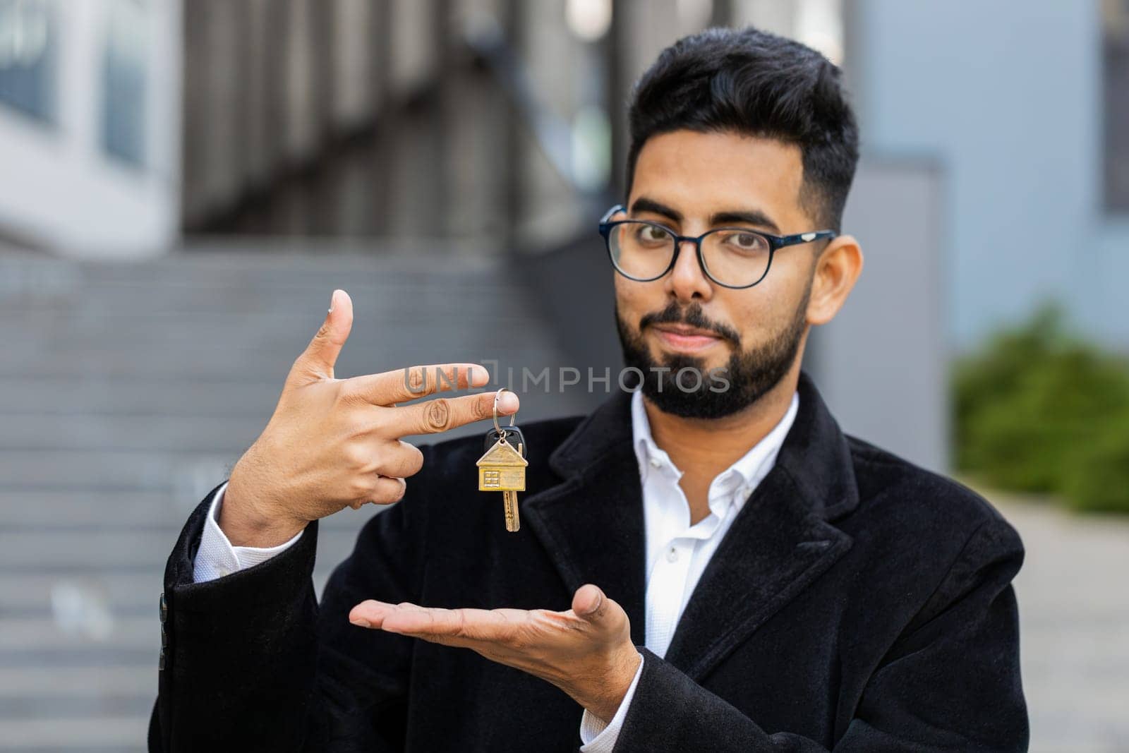 Indian businessman real estate agent lifting hand showing keys of new home house apartment buying or renting property, mortgage loan, investment outdoors. Arabian Hindu freelancer guy in downtown city