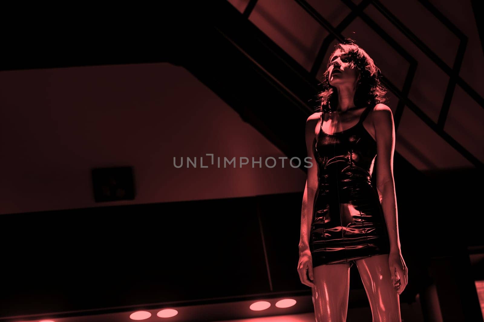A beautiful young sexy female model in latex stands in a red light in a hangar. Fetish concept