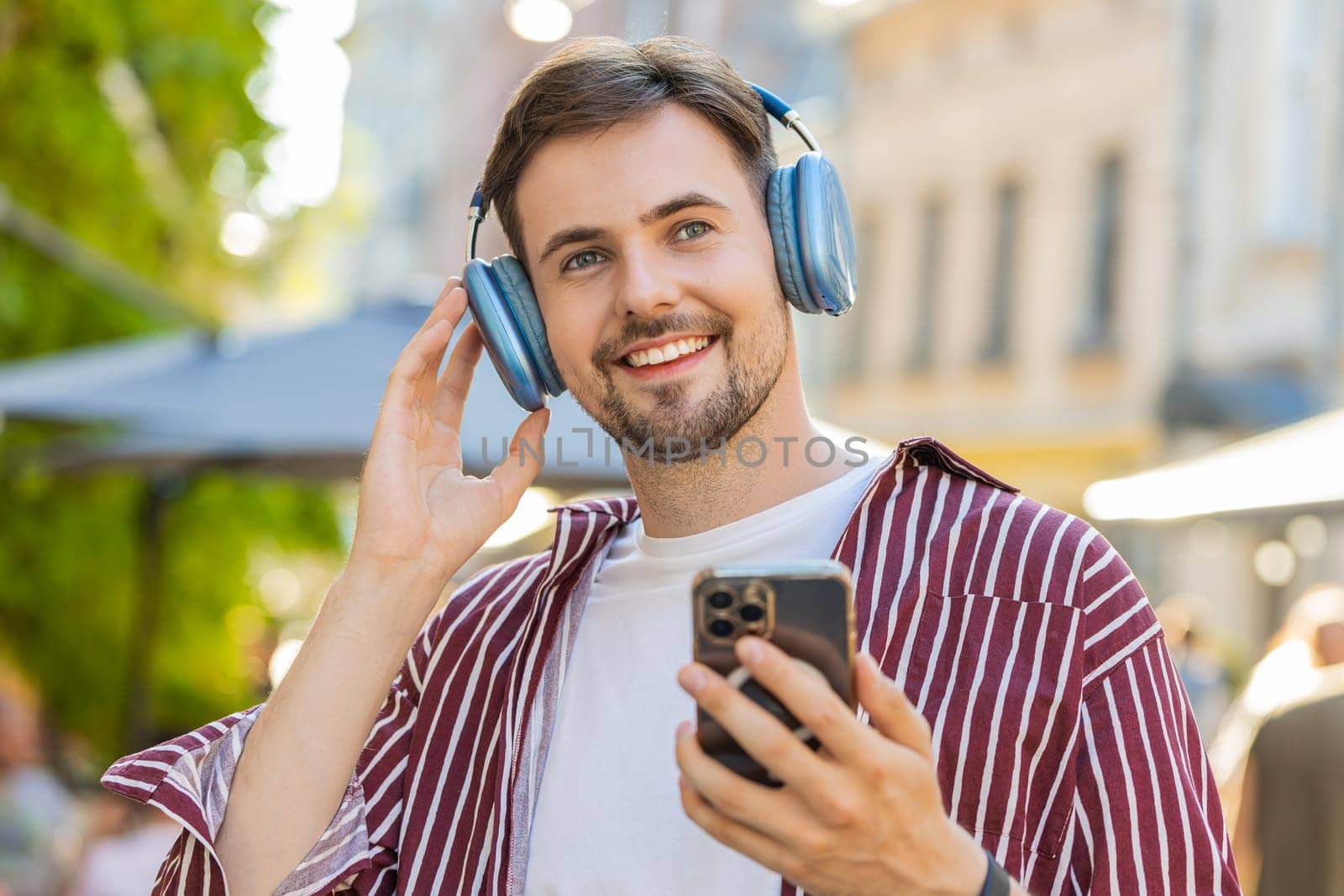 Happy relaxed overjoyed bearded adult man dancing in wireless headphones choosing, listening favorite energetic disco rock n roll music outdoors. Young guy standing on urban city sunshine street
