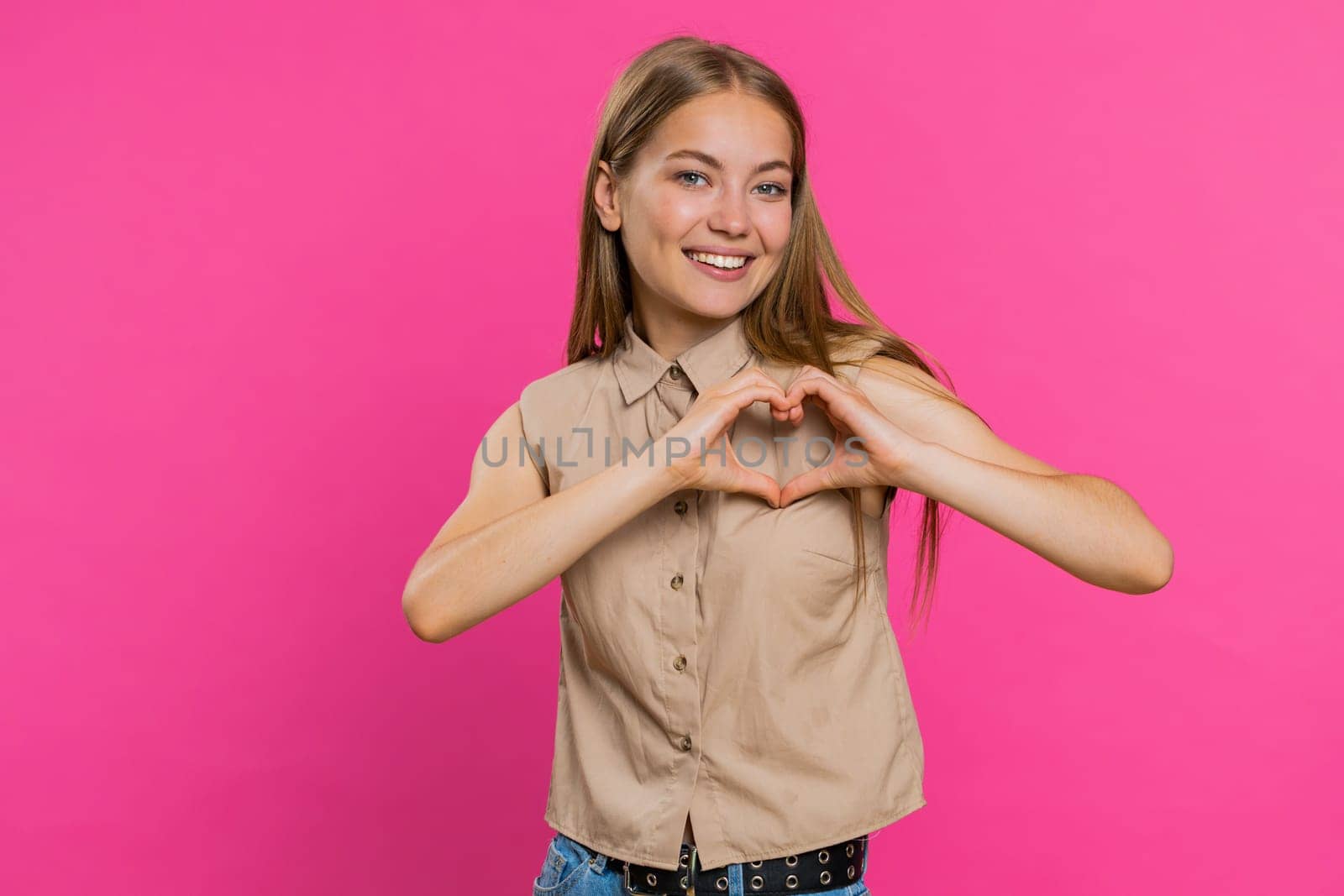 Woman in love. Smiling attractive woman makes heart gesture demonstrates love sign expresses good positive feelings and sympathy. Pretty blonde young girl isolated on pink studio wall background