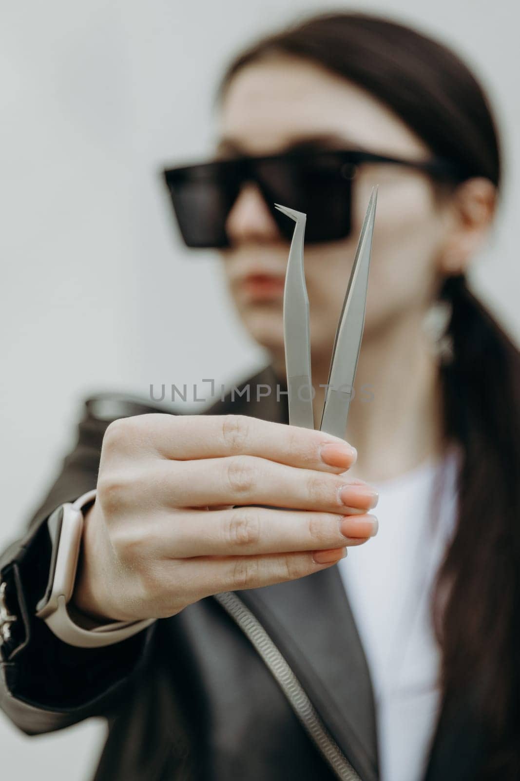 Portrait of one young beautiful Caucasian girl in sunglasses, leather jacket shows two eyelash extension tweezers with one hand, standing near a white wall on a spring day and looking to the side, close-up side view with depth of field.