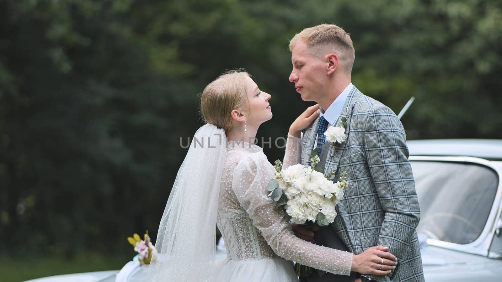 Newlyweds looking at each other in an embrace on their wedding day against a retro car backdrop. by DovidPro