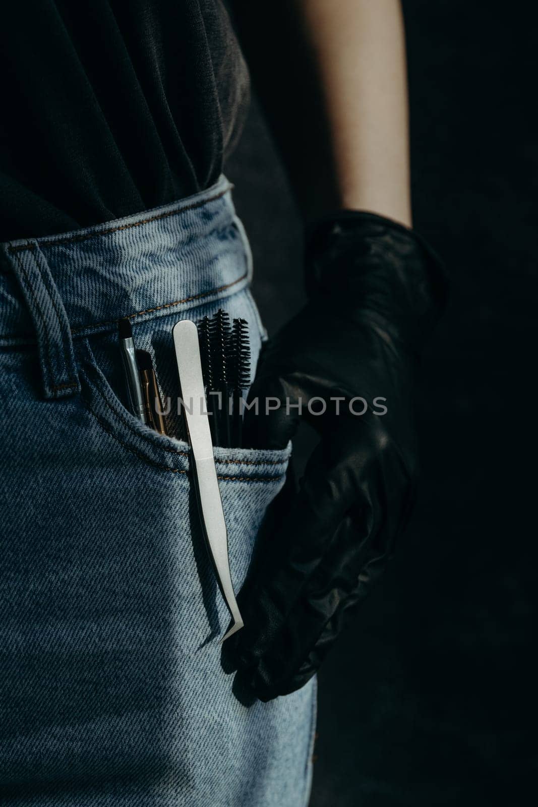 A set of brushes, tweezers and brushes for eyelash extensions stick out from the left pocket of blue jeans with a girl s hand in black gloves on a dark gray stone background, close-up side view. Dark style.