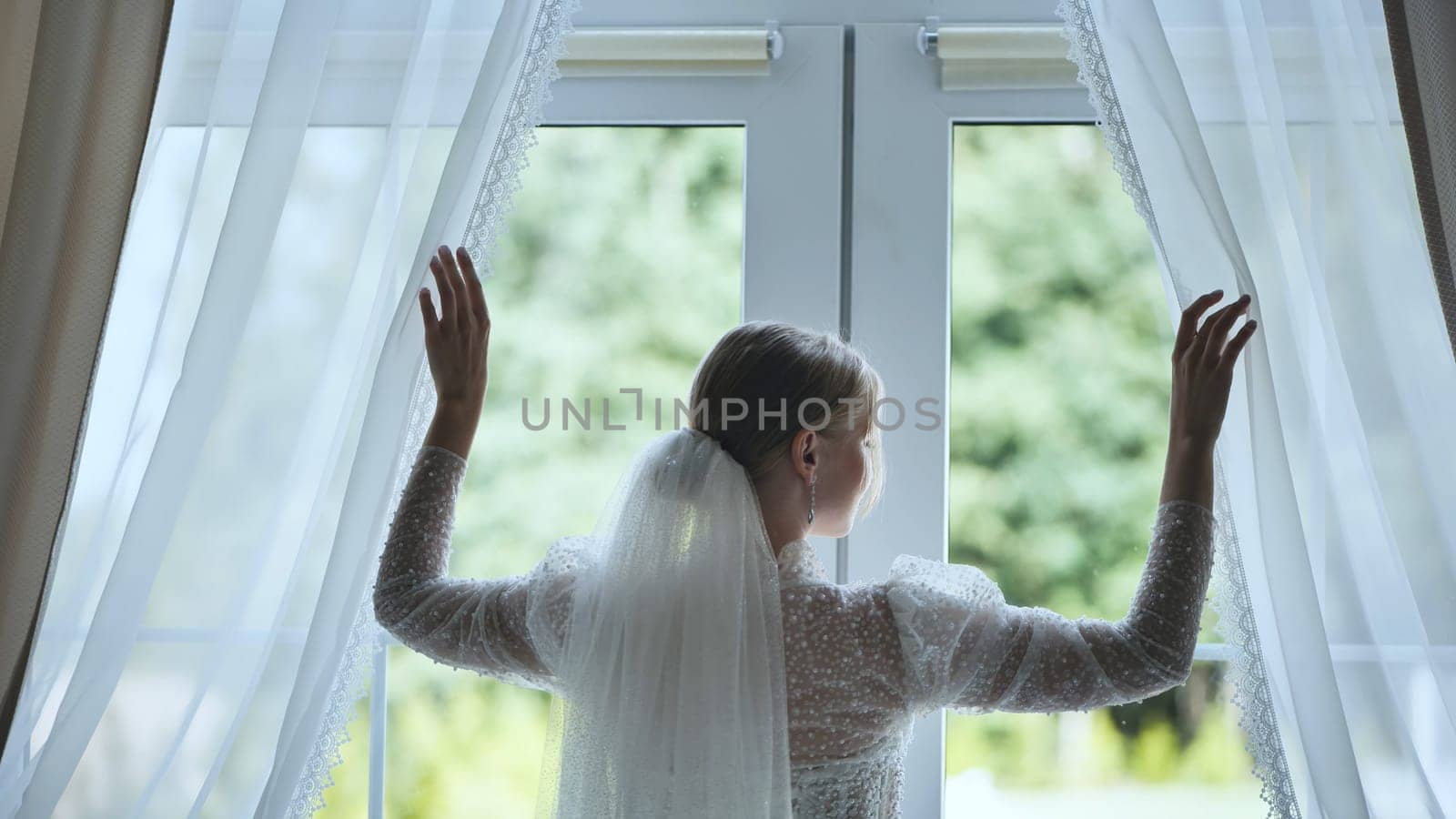 The bride adjusts the curtains at the window. by DovidPro