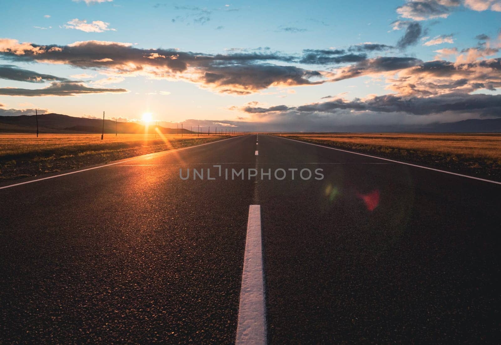 Empty intercity countryside asphalt road with the blue sky, sun and clouds on the background at sunset. Beautiful landscape