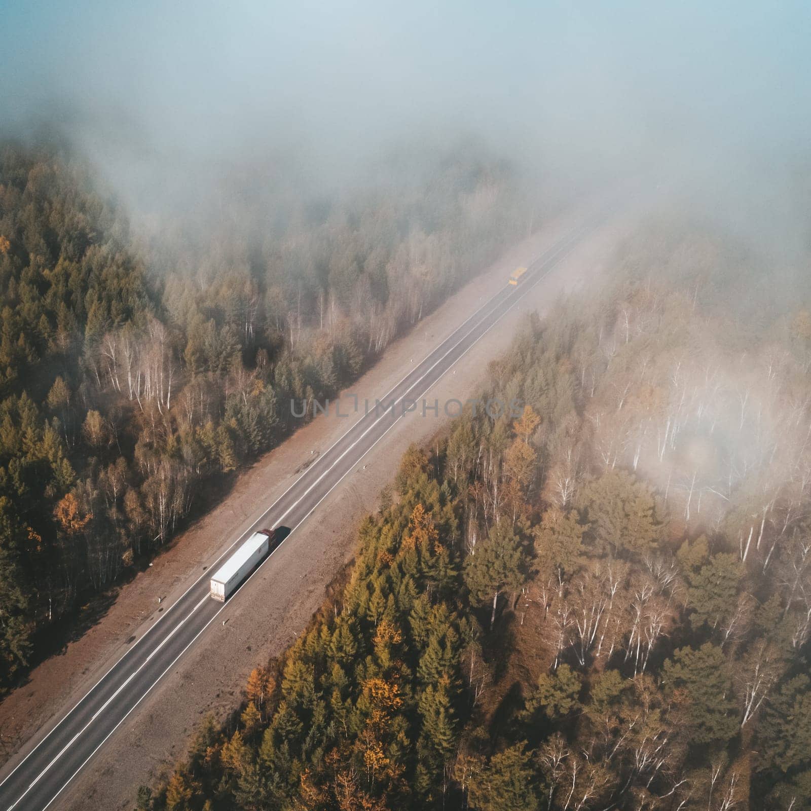Aerial shot of a lorry truck drives on an intercity road surrounded with autumn forest and with clouds above. Fall landscape by Busker