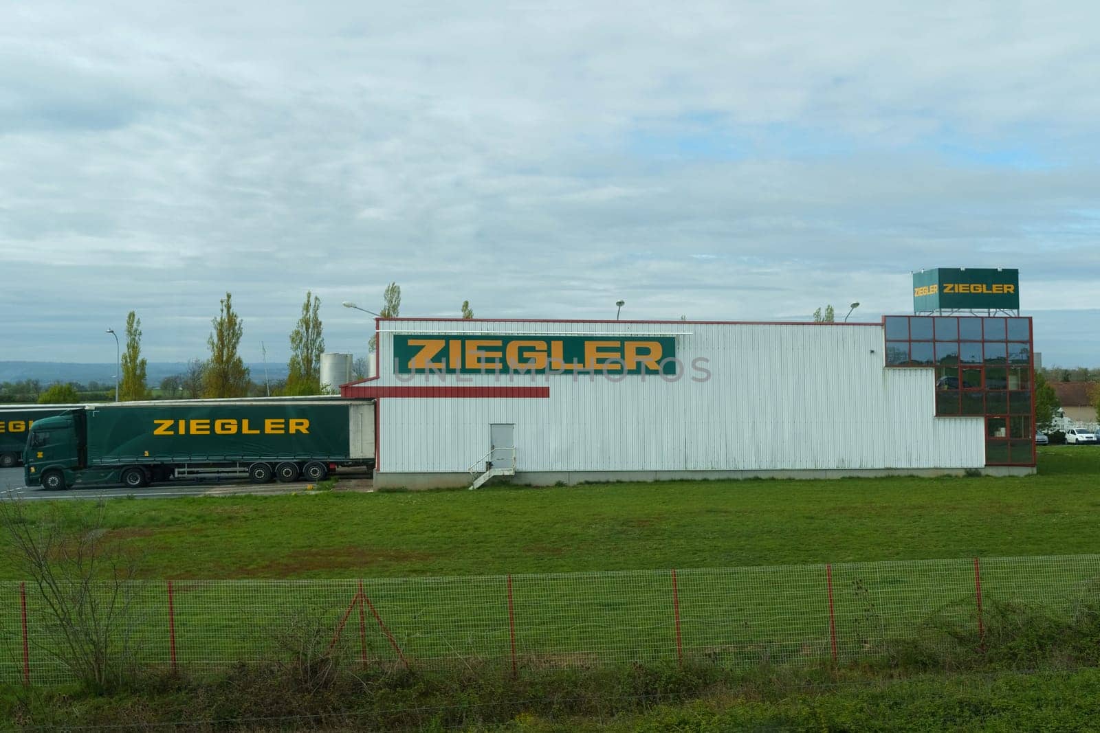 Clermont-Ferrand, France - April 26, 2023: A Ziegler logistics truck is parked outside a warehouse labeled with the Ziegler logo, with green fields and a cloudy sky in the backdrop.