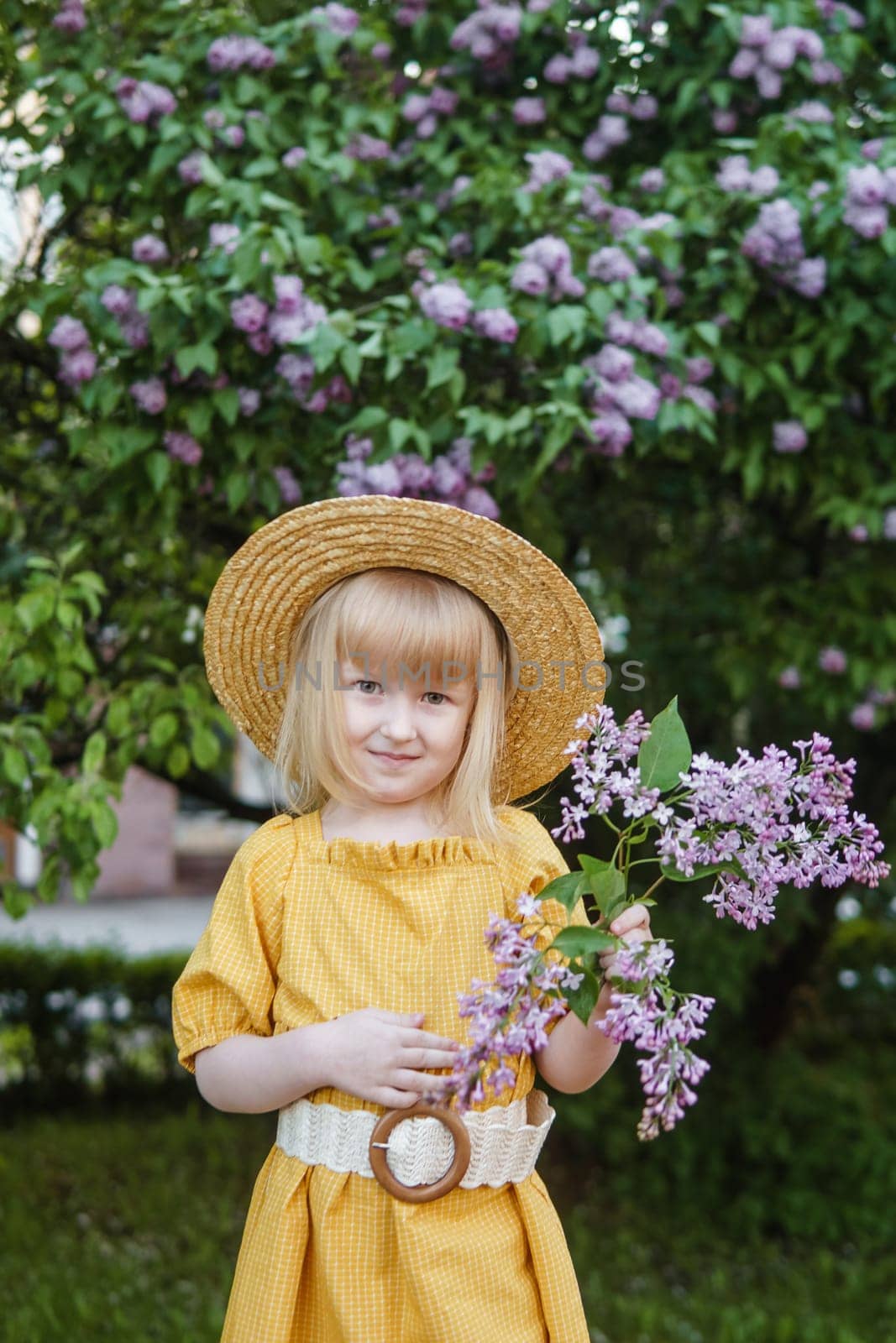 A little girl in a yellow dress and straw hat wearing a bouquet of lilacs. A walk in a spring park, blossoming lilacs