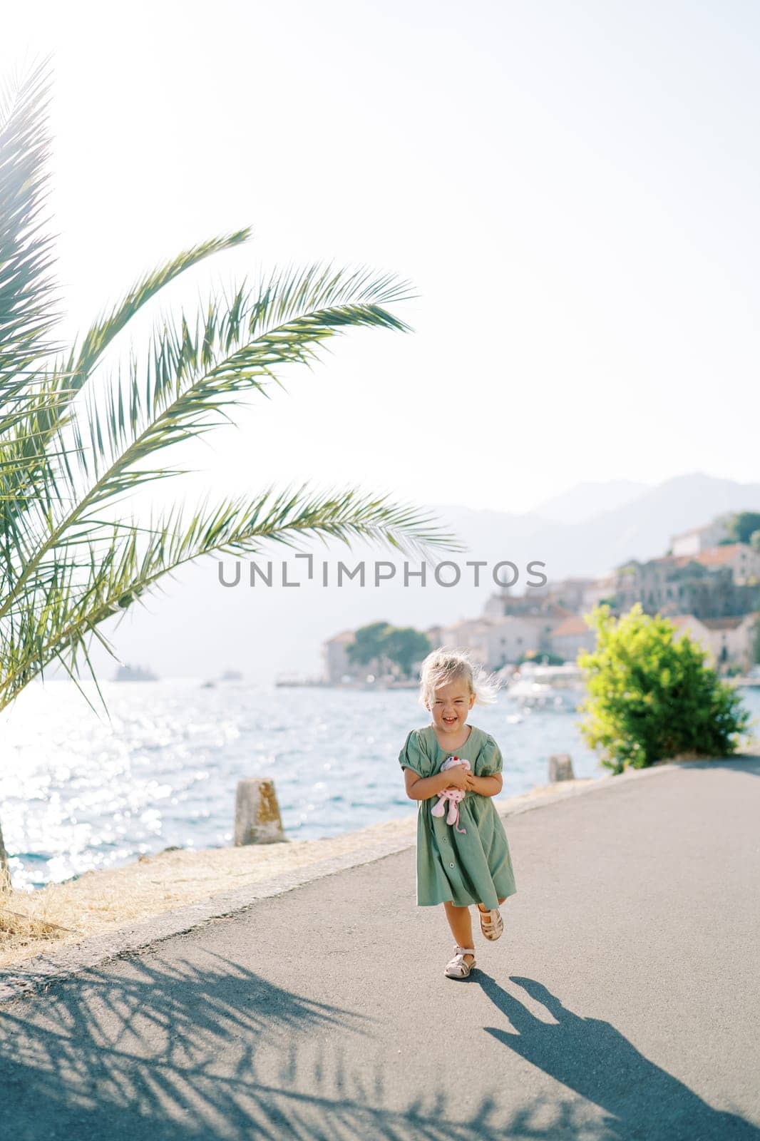 Little laughing girl with a toy walks along the embankment past a palm tree. High quality photo