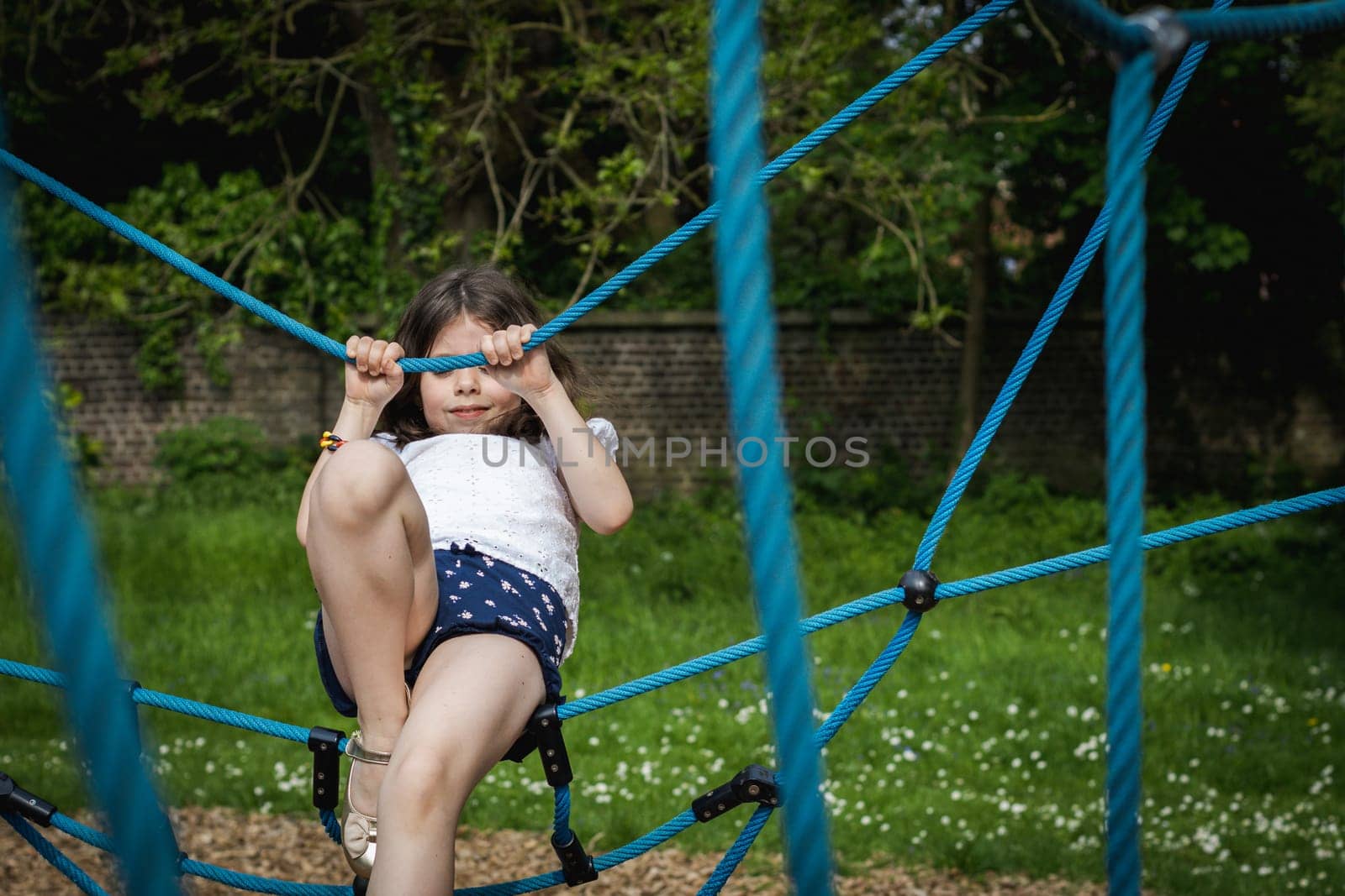 Portrait of one beautiful Caucasian happy brunette girl climbing on a blue rope swing on a playground on a summer day in a park on a playground, side view close-up.
