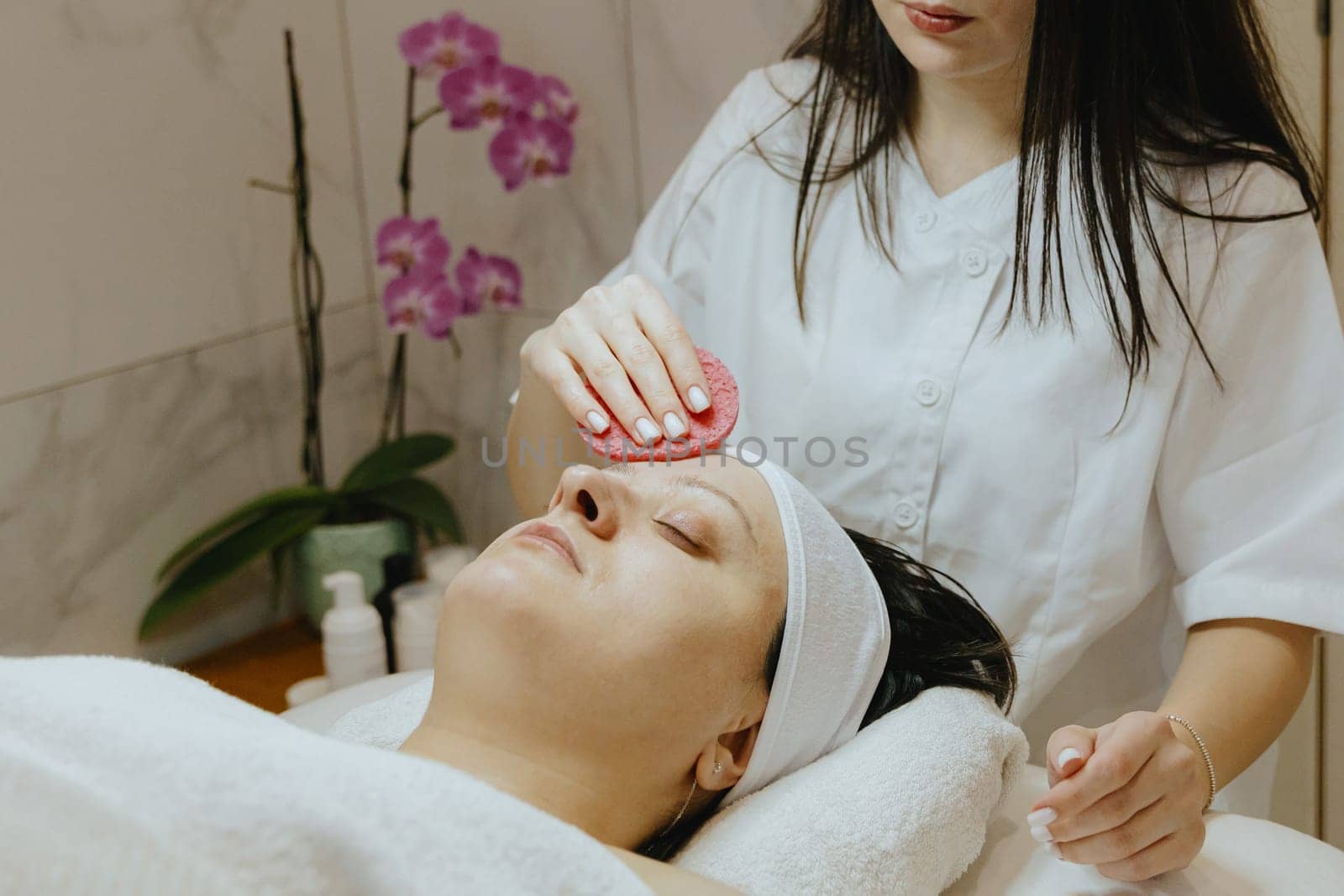 A young Caucasian girl cosmetologist cleanses the face of an adult female client using a pink scrubbing sponge, which lies on a massage table in a beauty salon, close-up side view.