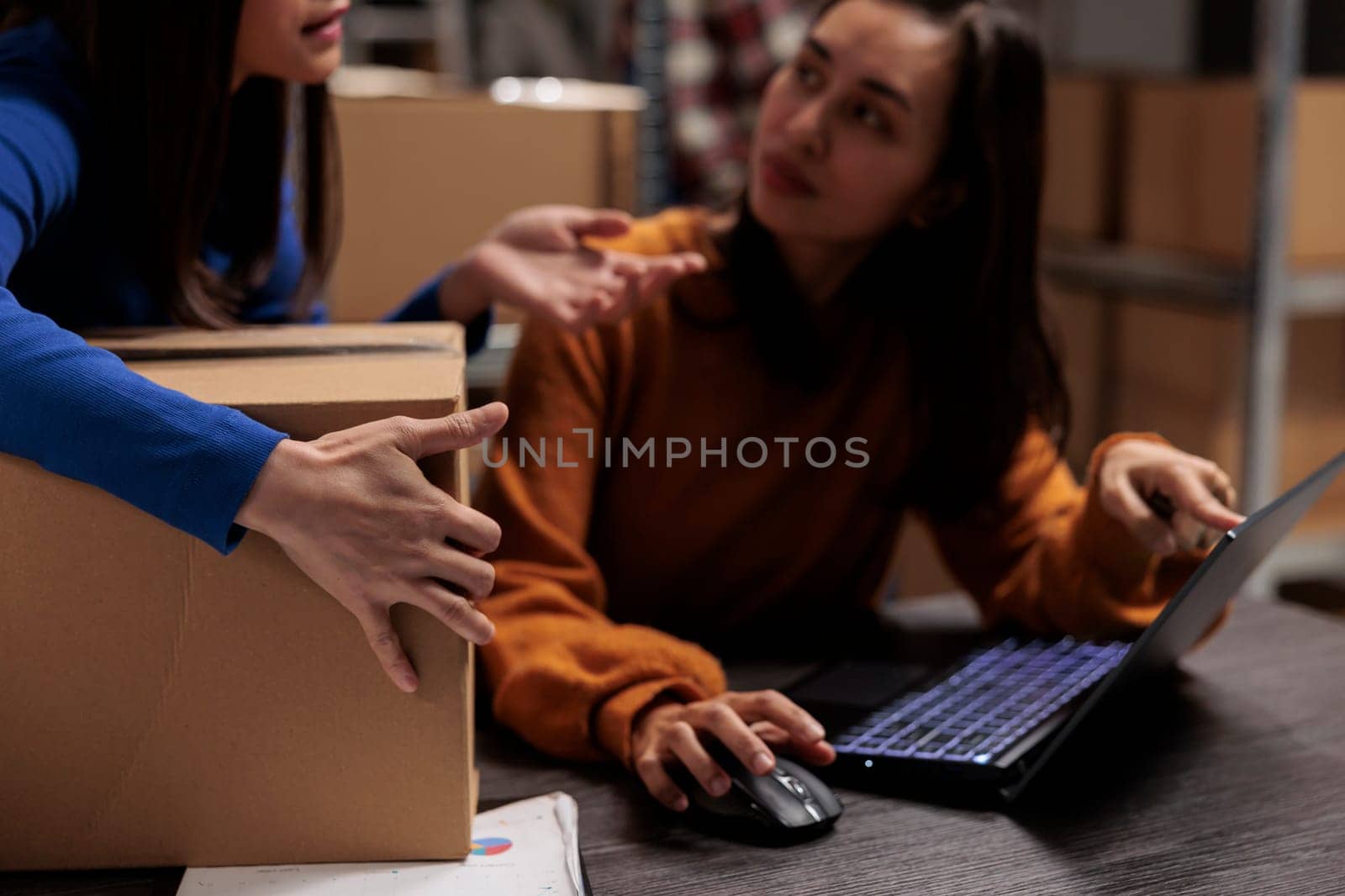 Storehouse employee asking coworker about pick ticket on laptop while working at desk in ecommerce retail business storage room. Colleagues tracking parcel shipment and preparing box for dispatching