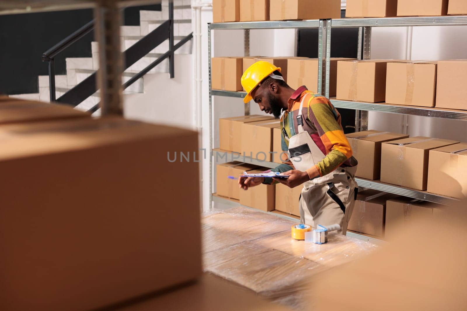 Storehouse supervisor analyzing merchandise checklist, standing beside cardboard boxes in storage room. African american manager wearing helmet and industrial overall working in warehouse