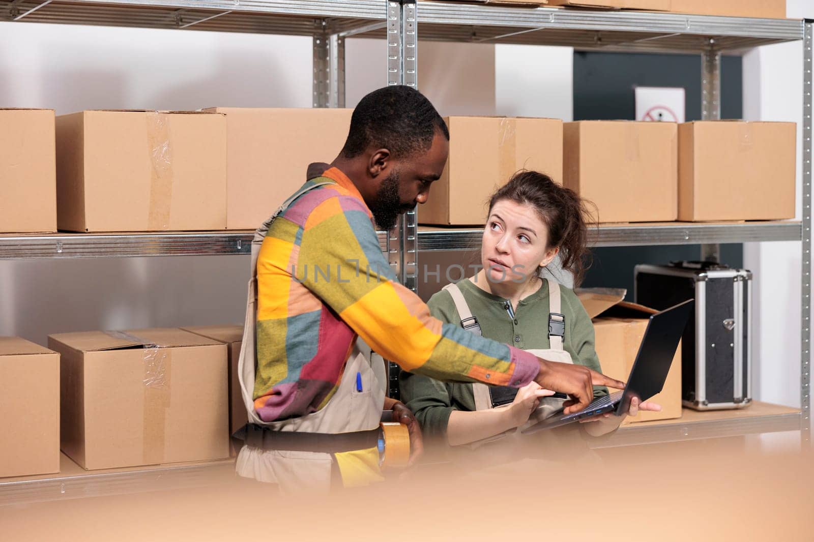 Multi ethnic stockroom supervisor packing parcels and managing delivery operations. Colleagues working in post office storage, standing beside cardboard boxes shelves preparing orders in storage room