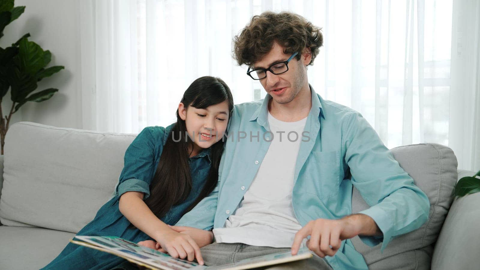 Happy daughter lean on father shoulder while listening story telling. Young asian schoolgirl looking ar dad while reading story book together and sitting at sofa. Family recreation concept. Pedagogy.