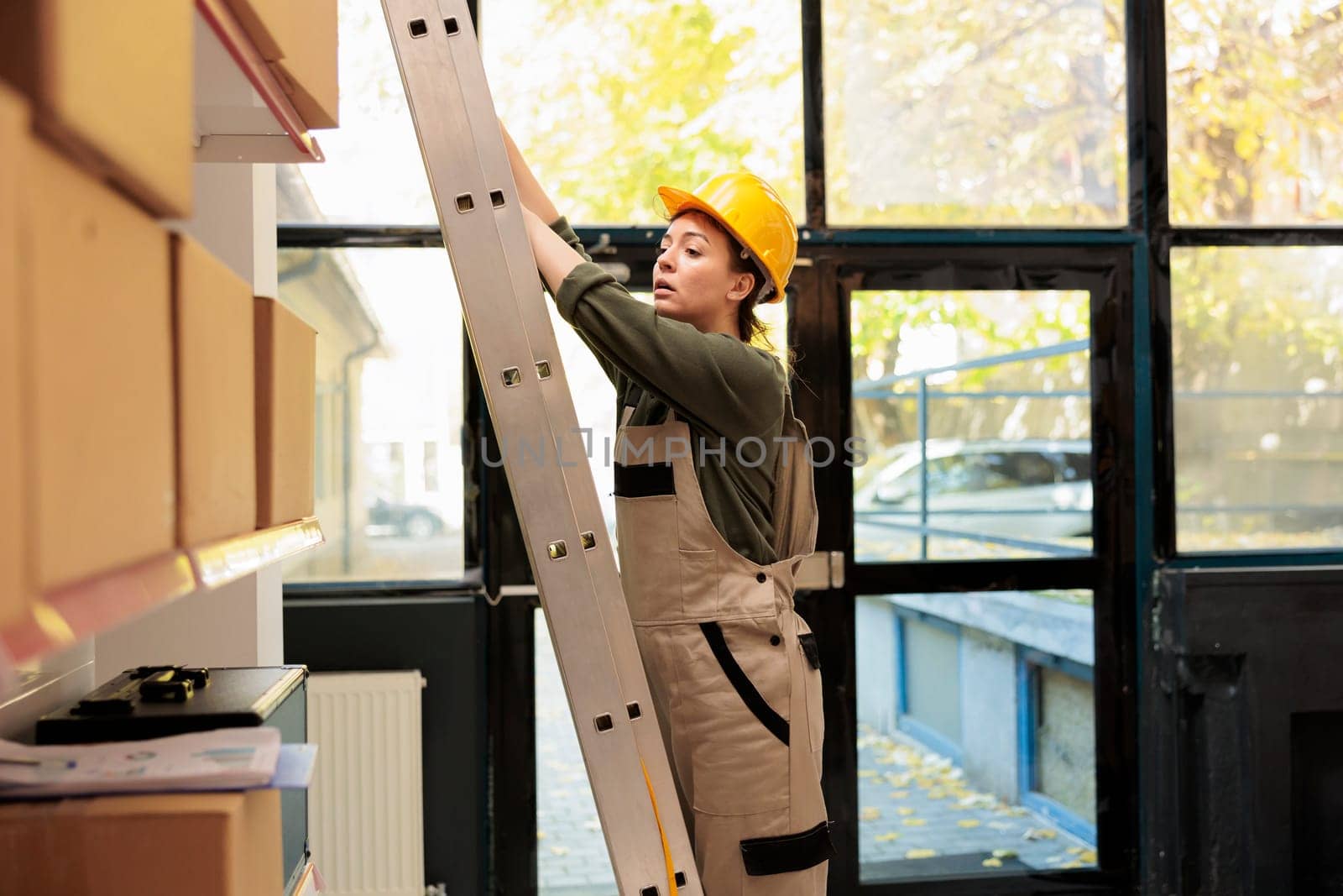 Storage room manager standing on ladder in warehouse, checking cardboard boxes full with merchandise. Supervisor woman working at customers orders before preparing delivery in storehouse