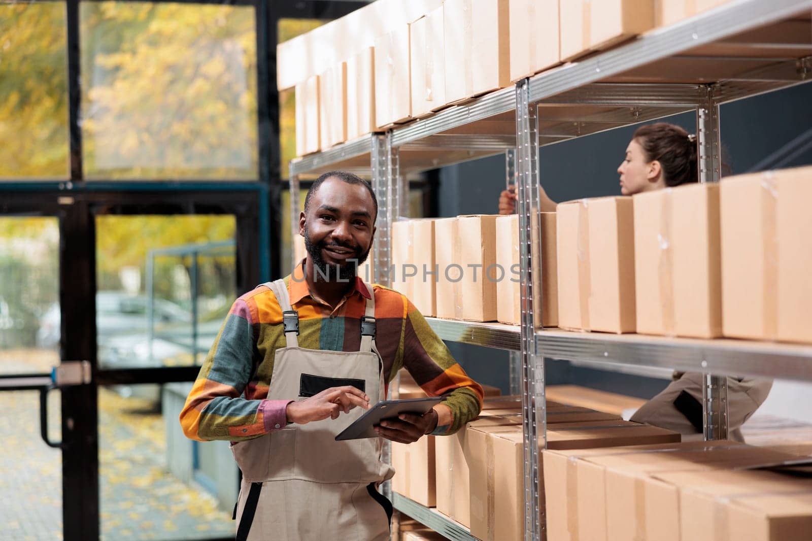 African american manager working at goods inventory, checking products on tablet computer in warehouse. Stockroom employee preparing customers packages, using cardboard boxes for delivery