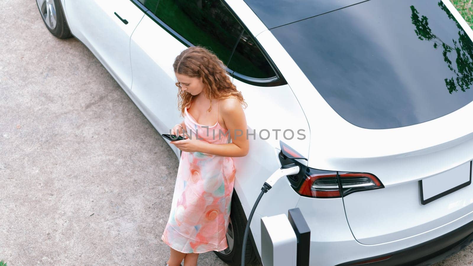 Aerial top view eco-friendly woman recharge electric vehicle from EV charging station, using EV technology utilization for tracking energy usage to optimize battery charging on smartphone. Synchronos