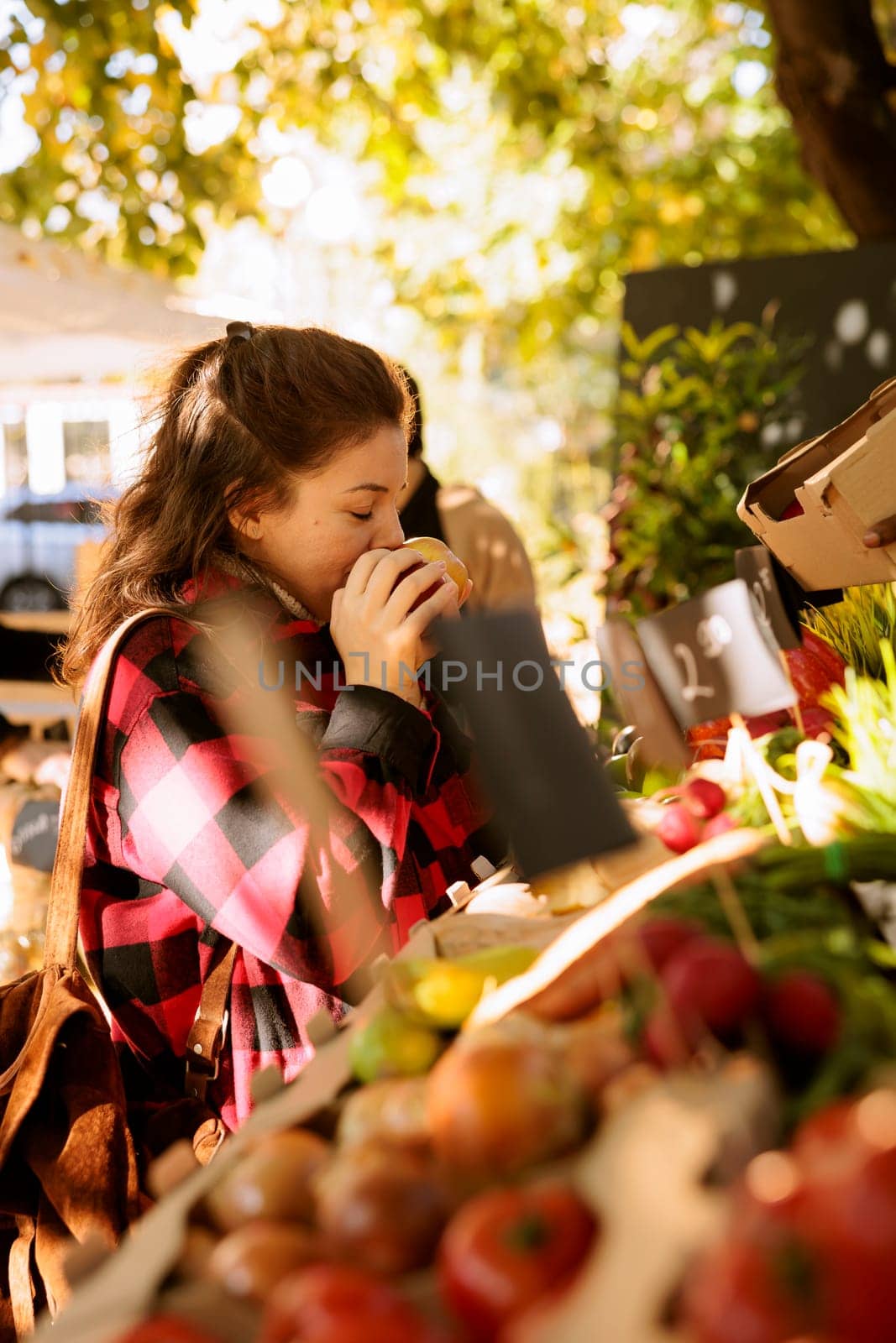 Selective focus of young adult smelling freshly harvested apple at eco friendly marketplace. Female shopper choosing and purchasing locally grown organic bio fruits and veggies at outdoor food market.