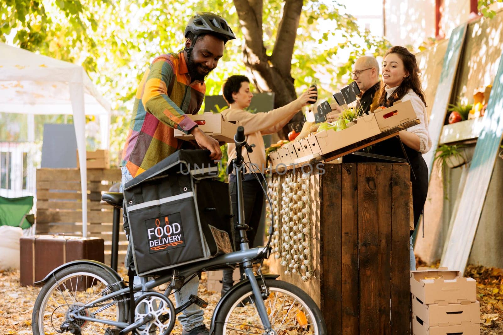 Black male courier putting box of organic products in backpack for delivering order to clients, using bicycle. Young cheerful deliveryman working on farmers market veggies shipping.