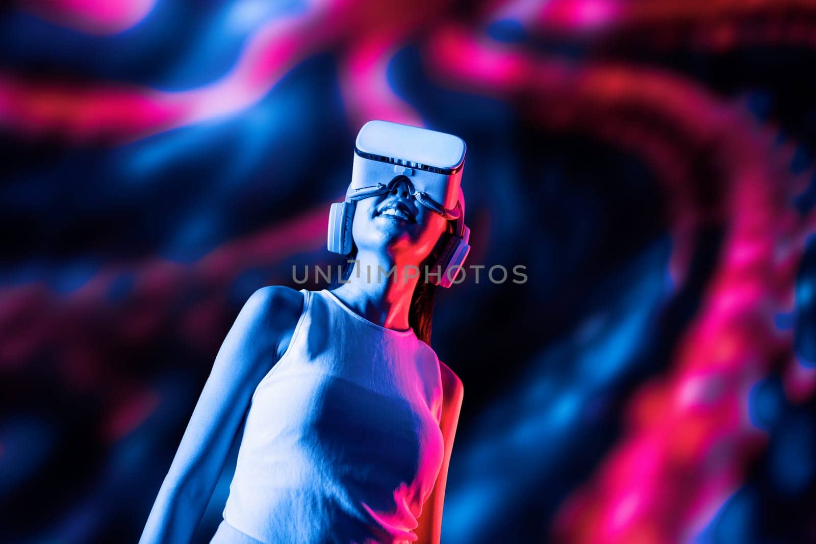 Smart female standing surrounded by neon light wearing VR headset connecting metaverse, future cyberspace community technology. Elegant woman looking faraway and smiling satisfactorily. Hallucination.