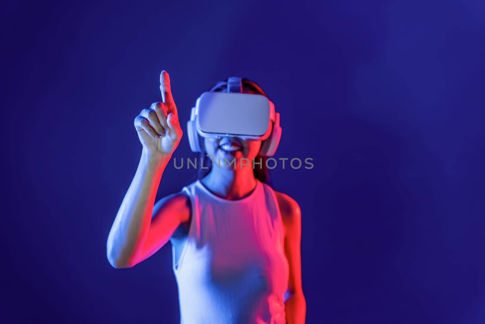 Smart female stand with surrounded by cyberpunk neon light wear VR headset connecting metaverse, futuristic cyberspace community technology. Woman using finger pointing virtual object. Hallucination.