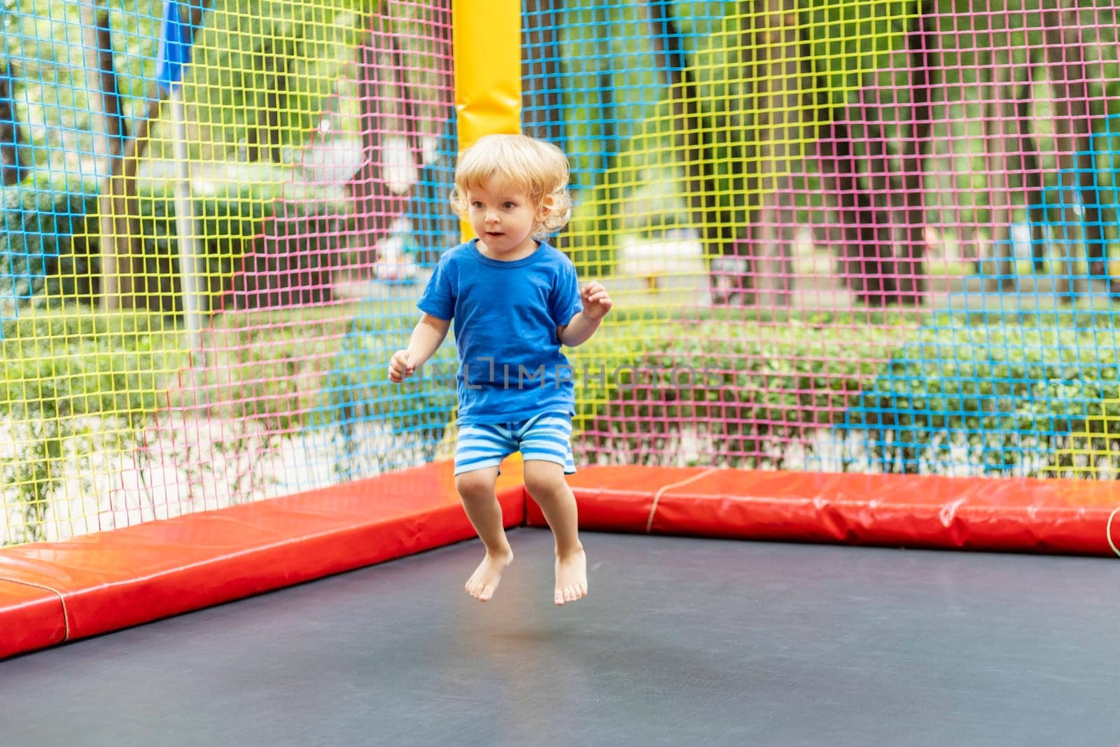 Toddler boy in blue outfit jumping on colorful outdoor trampoline. Playful activity and fun concept. Design for banner, poster.