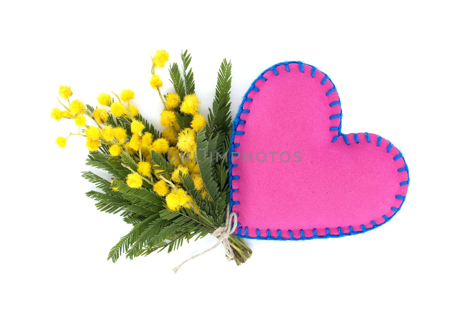 Heart-shaped pink pillow beside of yellow mimosa flowers against a white background, representation of a heartfelt sentiment expressed through the language of colors and shapes