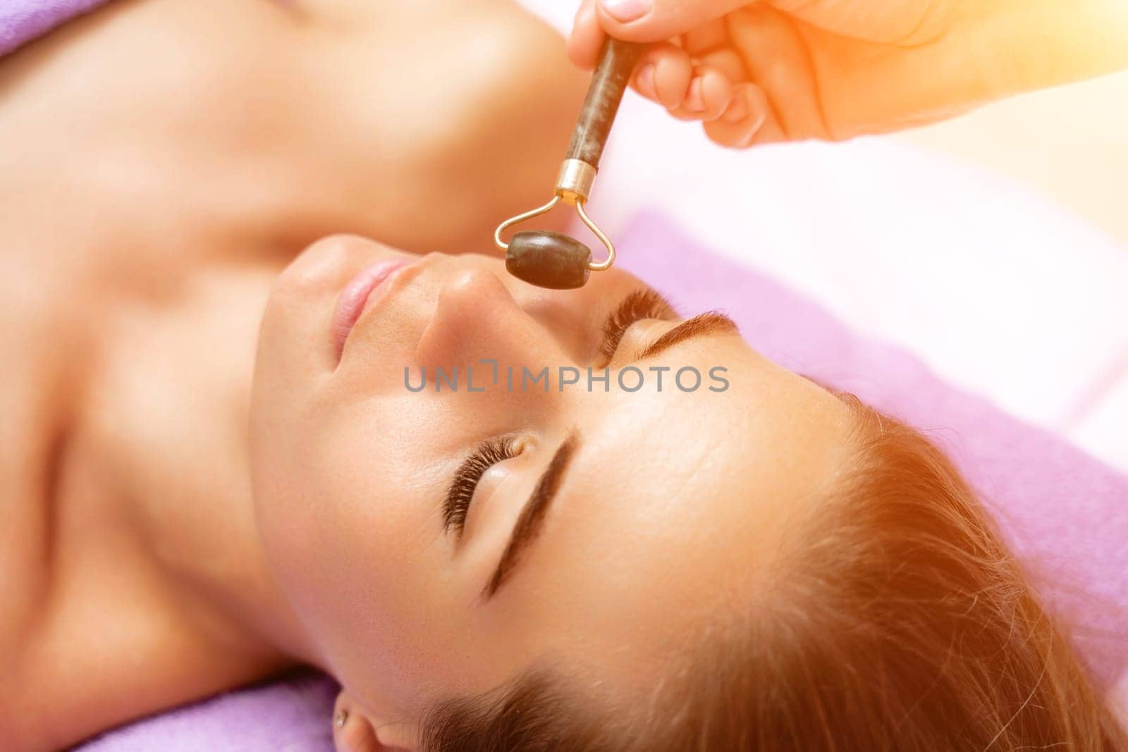 Relaxing massage. Side view european woman getting massage with jade face roller gouache in spa salon by Matiunina