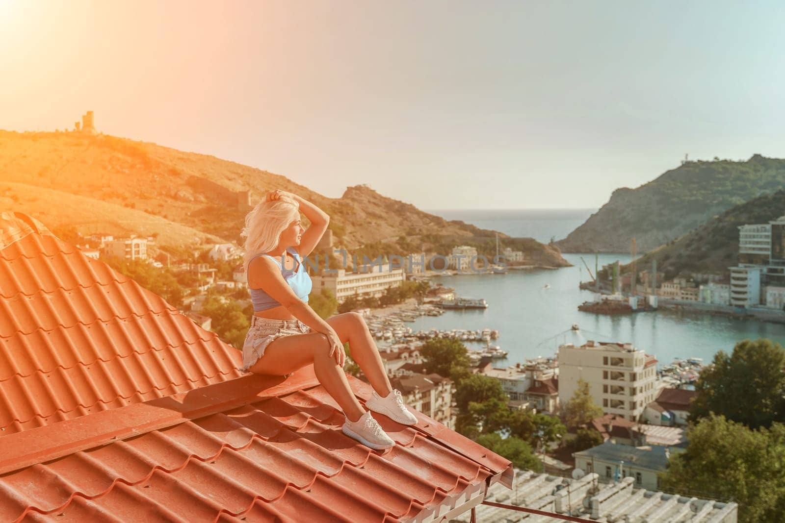 Woman sits on rooftop, enjoys town view and sea mountains. Peaceful rooftop relaxation. Below her, there is a town with several boats visible in the water. Rooftop vantage point. by Matiunina