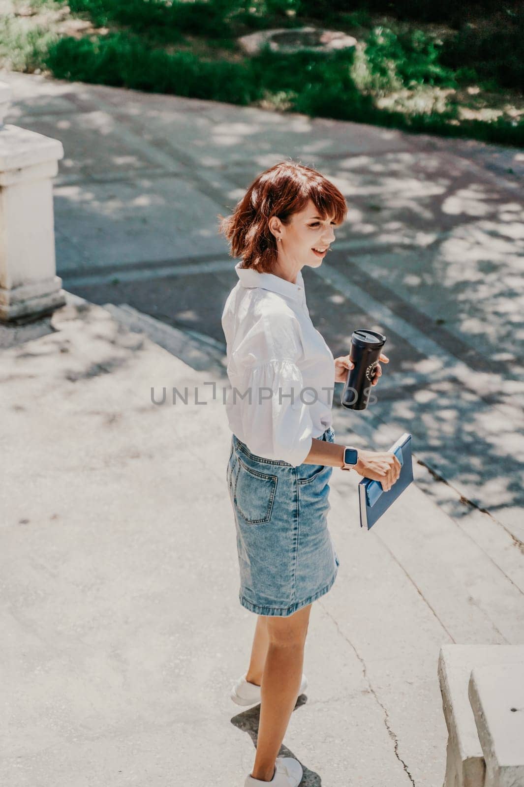 Business woman city street. A woman in a white shirt and blue denim skirt walks down the street holding a blue notebook in her hands. Summer city walks, travel by Matiunina
