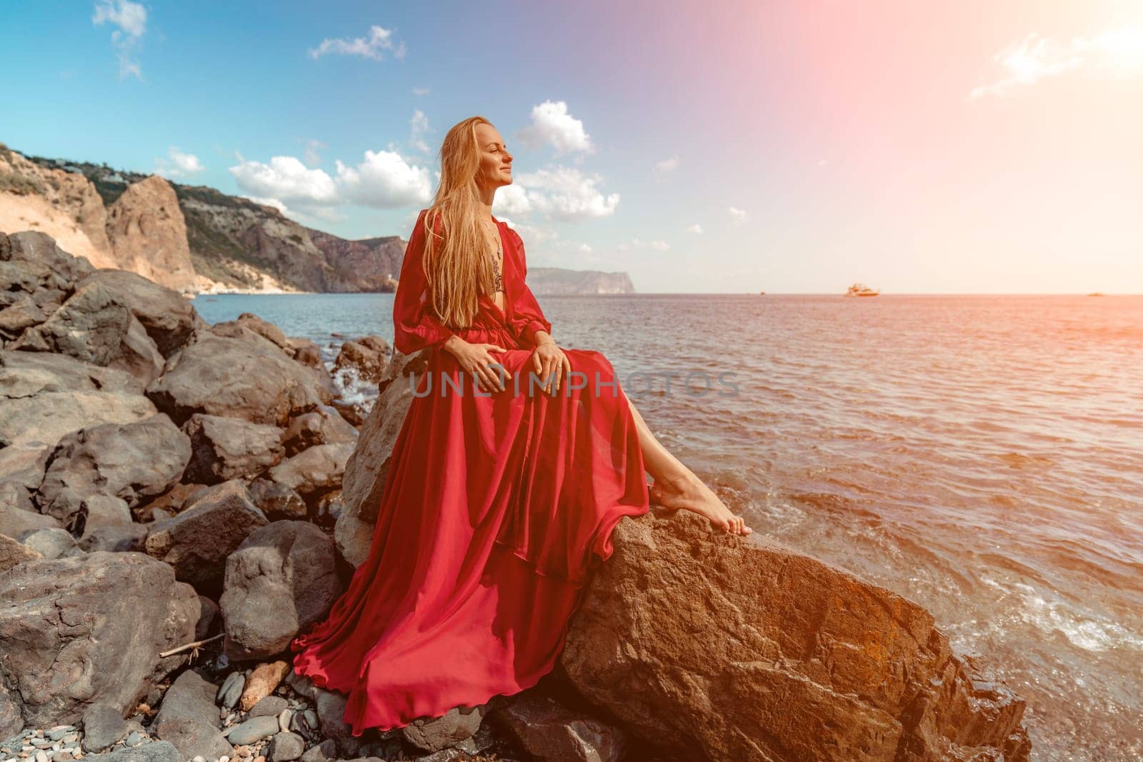 Red dress sea woman. A blonde with flowing hair in a long red dress sits on a rock near the sea. The concept of trips, a photo shoot at the sea by Matiunina