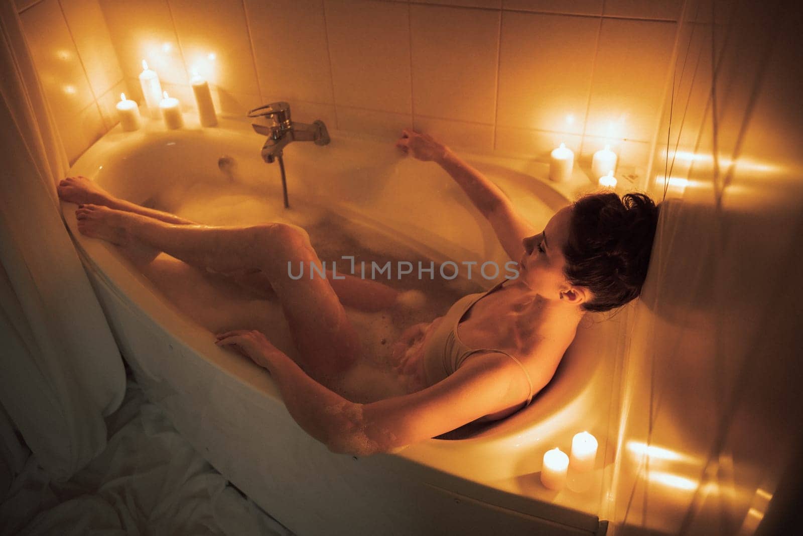 A woman is sitting in a bathtub with candles lit around her. The candles are lit in a way that they create a warm and relaxing atmosphere