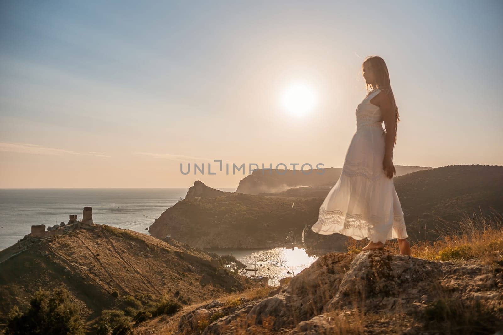 A woman stands on a rocky hill overlooking the ocean. She is wearing a white dress and she is enjoying the view. The scene is serene and peaceful, with the sun shining brightly in the background