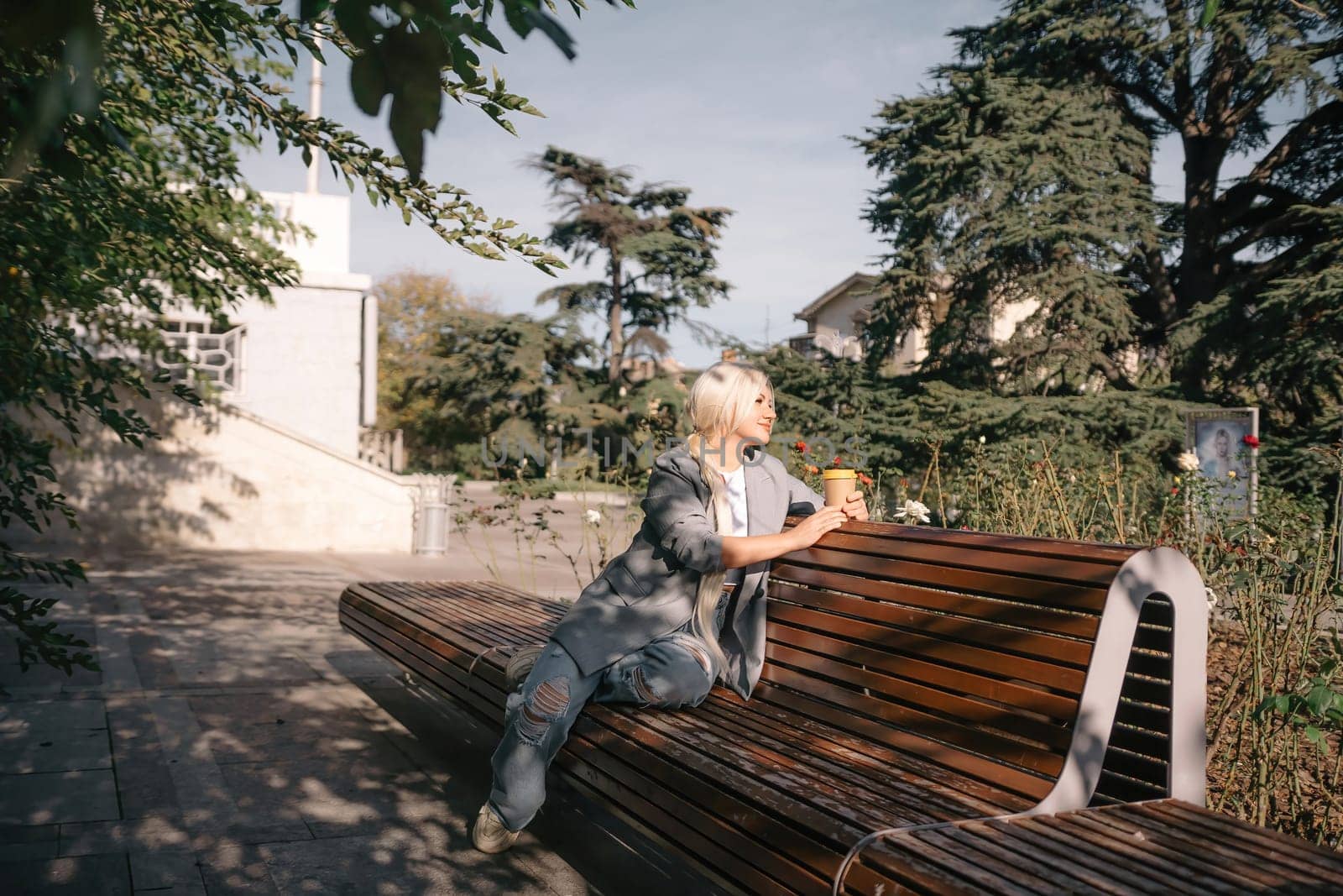 A blonde woman sits on a bench with a cup of coffee in her hand. She is wearing a gray jacket and jeans. The bench is wooden and has a curved back. The scene is peaceful and relaxing